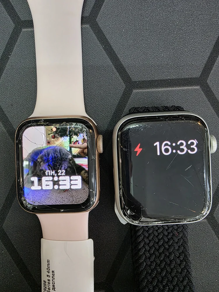 Двое Apple Watch с разбитыми стёклами лежат рядом на чёрном коврике мастерской, обе матрицы работают: на одних часах видно время 16:33 и красный значок низкого заряда, на других под трещинами читается фотография на циферблате