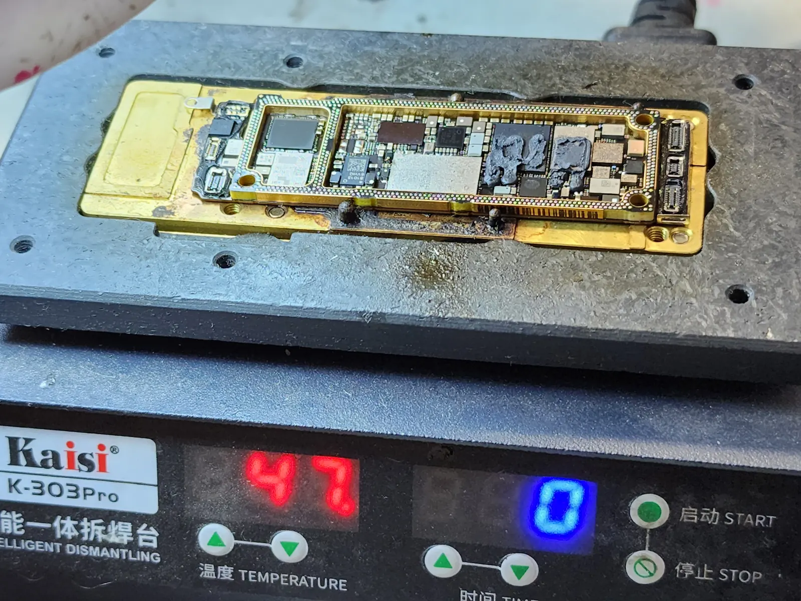 Плата iPhone из двух половинок лежит в нагревательном столе Kaisi K-303Pro; верхнюю половинку уже сняли, видны микросхемы под тёмным компаундом, красное табло показывает 47.