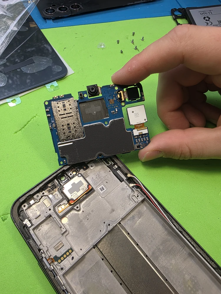 В руке мастера снятая плата Android-смартфона с камерами и сканером отпечатка над пустой рамкой корпуса; рядом по зелёному коврику разложены винты и снятые детали.