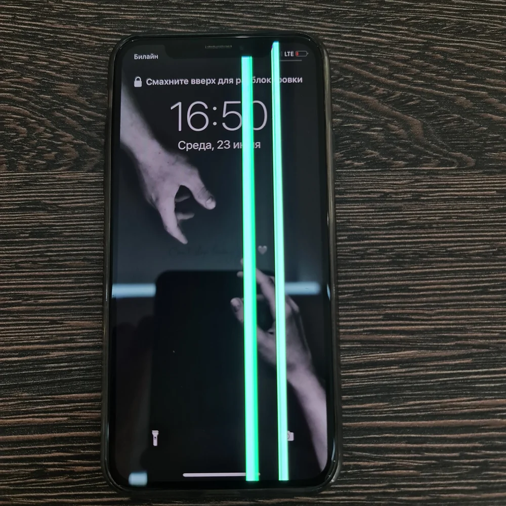 iPhone лежит экраном вверх у края тёмного деревянного стола, поверх экрана блокировки с часами 16:50 сверху донизу идут две яркие зелёные вертикальные линии.