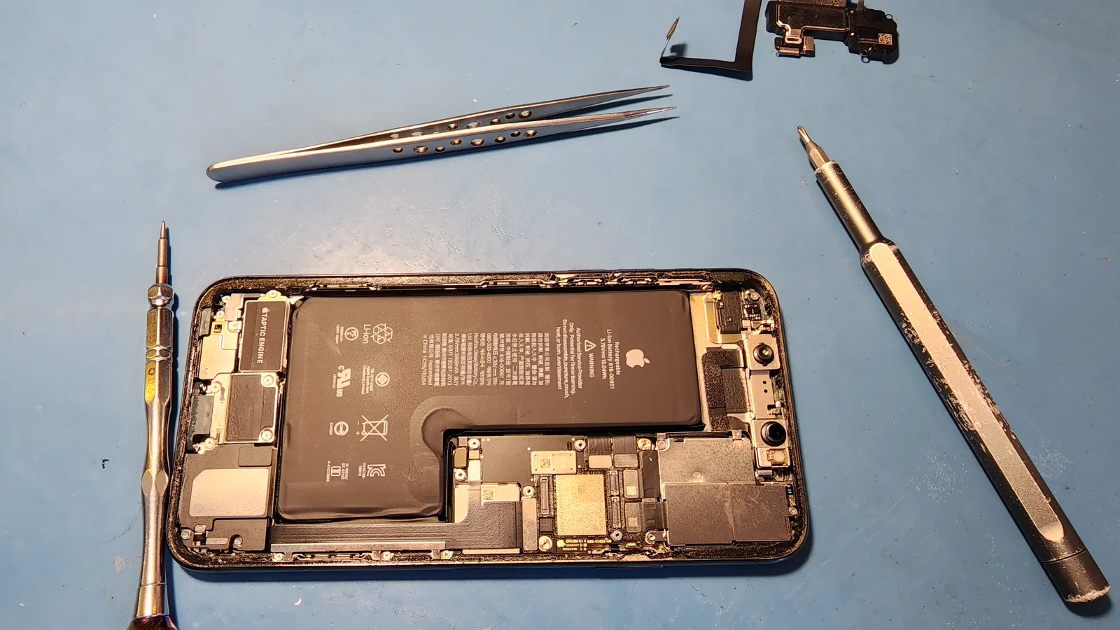 Разобранный iPhone на столе мастера: снятая крышка, аккумулятор, системная плата, динамик и вибромотор, рядом отвёртки и пинцет
